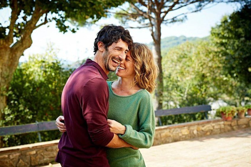 A couple embracing outdoors with trees and a stone wall in the background. 1