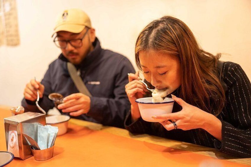 Two people sitting at a table with bowls of food, a tissue box, and a small metal container. 5