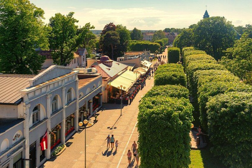 A street lined with buildings, a row of trees, and people walking. 2