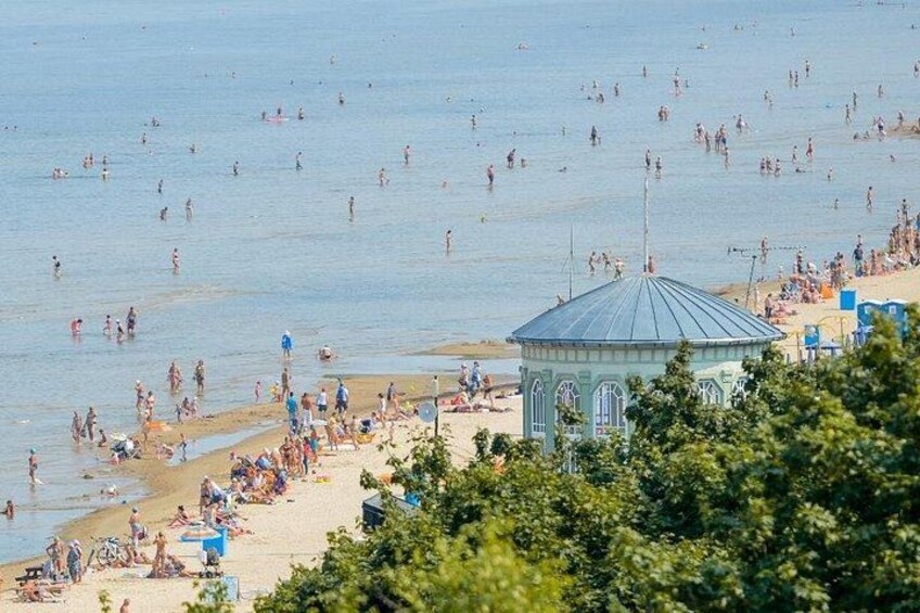 A crowded beach with people swimming and sunbathing, a gazebo-like structure, and dense foliage in the foreground. 3