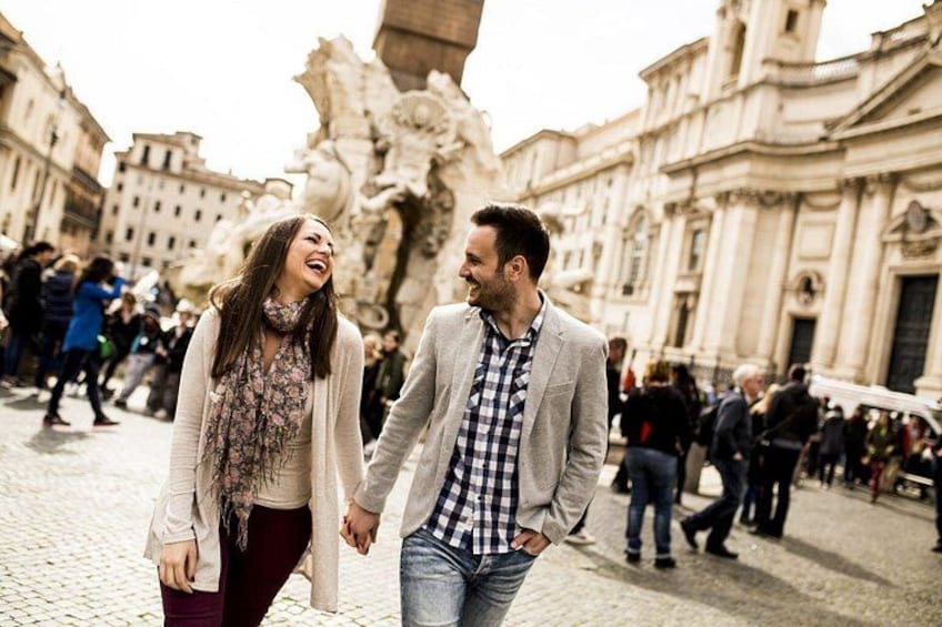 A couple walking hand in hand in a cobblestone square with historic architecture and a large fountain in the background. 2