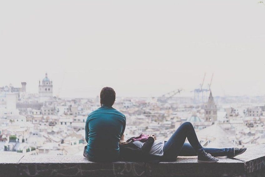 A person sitting on a ledge overlooking a cityscape with buildings and a prominent tower. 1