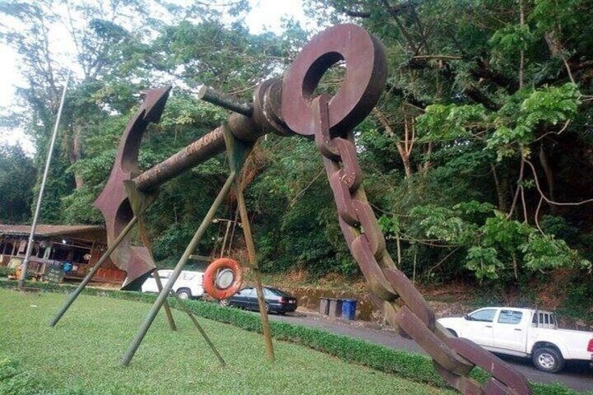 A large metal sculpture resembling a figure with a circular head and a long body, set in a grassy area with trees and a parked car in the background. 2