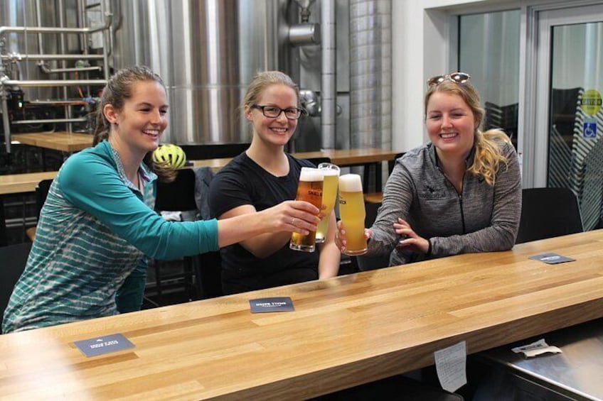 Trois personnes trinquent avec des verres de bière dans un bar. On aperçoit un comptoir en bois et des cuves métalliques à l’arrière-plan. 4