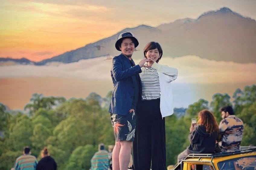 A couple standing with a scenic mountain backdrop, a vehicle, and people in the distance. 1