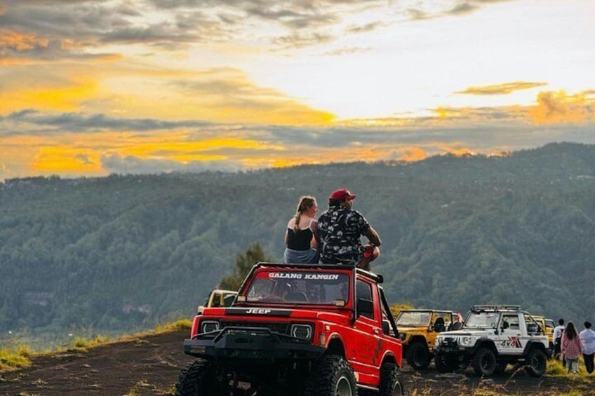 A red Jeep with a person standing on top, a white vehicle in the background, and a mountainous landscape with a sunset. 5