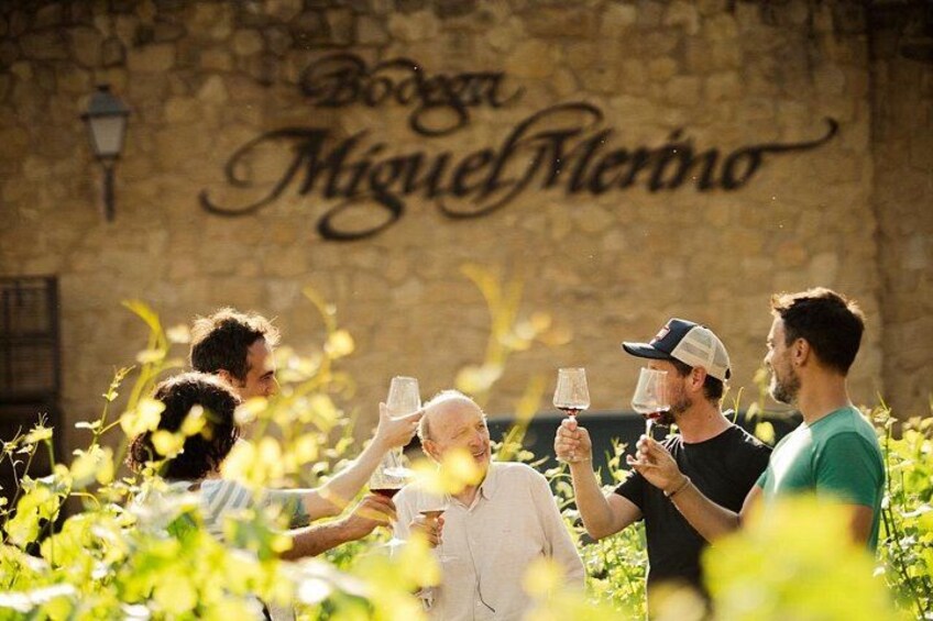 A group of people toasting with wine glasses in a vineyard with a stone wall and a sign that reads 'Bodega Miguel Merino'. 1