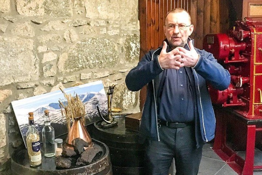 A man in a blue jacket standing in front of a rustic stone wall with a vintage red boiler and a painting of a mountain landscape. 5