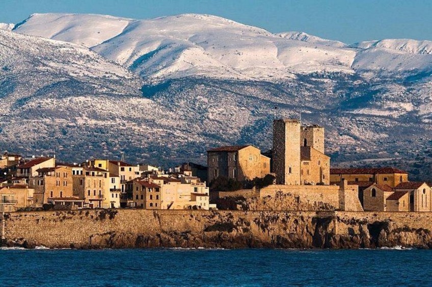 A coastal town with historic buildings, a prominent tower, and snow-covered mountains in the background. 1