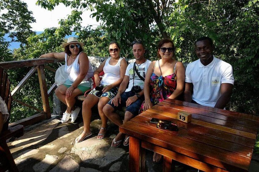 A group of people sitting on a wooden bench outdoors with greenery in the background. 4