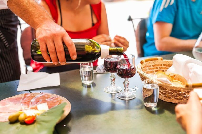A person pouring wine into a glass, a plate with food, and a basket with bread. 2