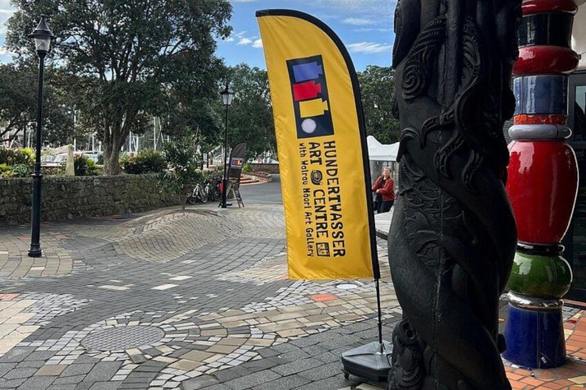 Una bandiera gialla con il testo 'HUNDERTWASSER ART CENTRE', un muro di pietra, un pavimento decorato e una statua. 5
