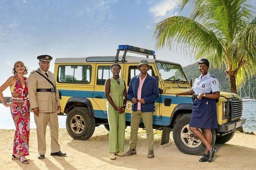A group of people standing in front of a yellow and blue vehicle on a sandy beach with palm trees and a body of water in the background. 1