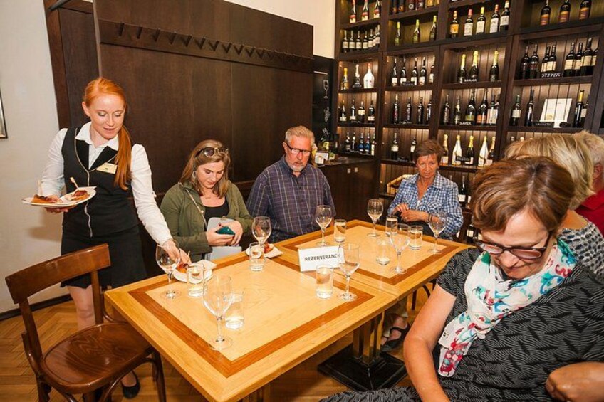 A wine tasting event with guests seated at tables, a waiter serving plates, and a wine rack in the background. 4
