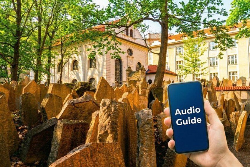 Una mano sostiene un teléfono inteligente con la etiqueta 'Audio Guide', frente a un muro de piedra y un edificio al fondo. 1