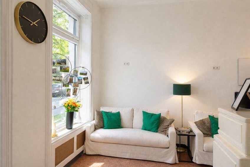 A cozy room with a white sofa, green cushions, a clock, a lamp, and a window with pictures. 2