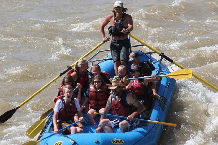 A group of people in a blue inflatable raft on a river, paddling with yellow paddles. 1