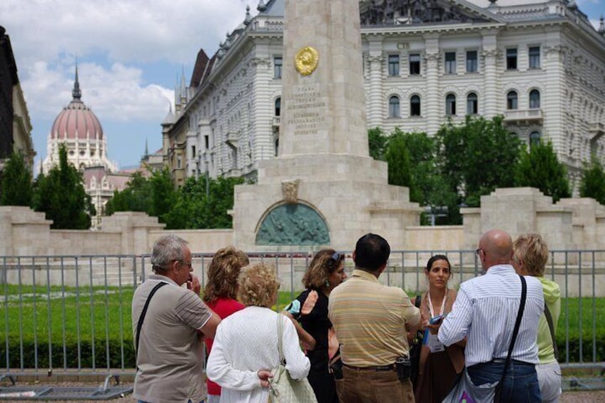 Un grupo de personas de pie frente a un monumento con un reloj, un gran edificio con una cúpula al fondo y una cerca rodeando el área. 3
