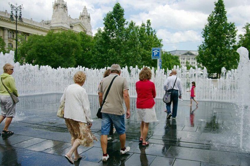 Un grupo de personas caminando cerca de una fuente en una plaza pública con un edificio histórico al fondo. 2