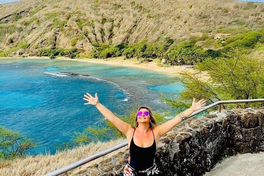 A person with arms outstretched overlooking a coastal landscape with clear blue waters and a sandy beach. 1