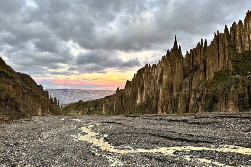 A rugged landscape with a narrow canyon flanked by tall, jagged rock formations under a cloudy sky. 1