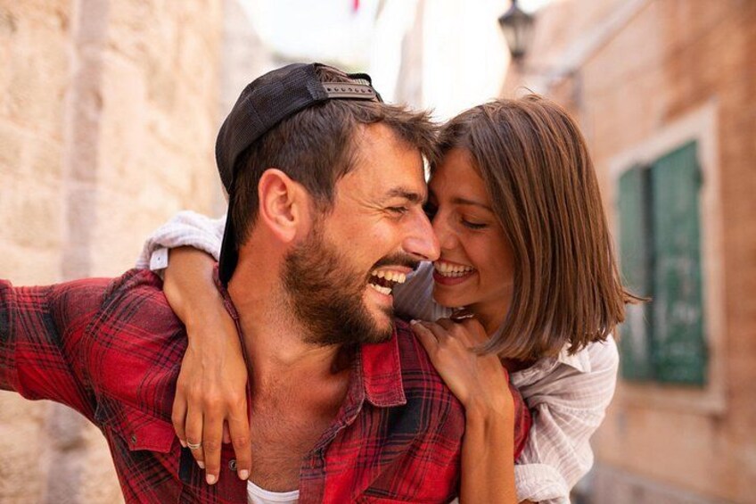 A person carrying another on their back, both wearing casual clothing, in a narrow street with stone buildings. 4