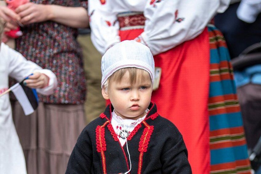 Un bambino con abiti tradizionali, un cappello bianco e un indumento nero con bordi rossi, in piedi tra adulti vestiti con costumi colorati. 5