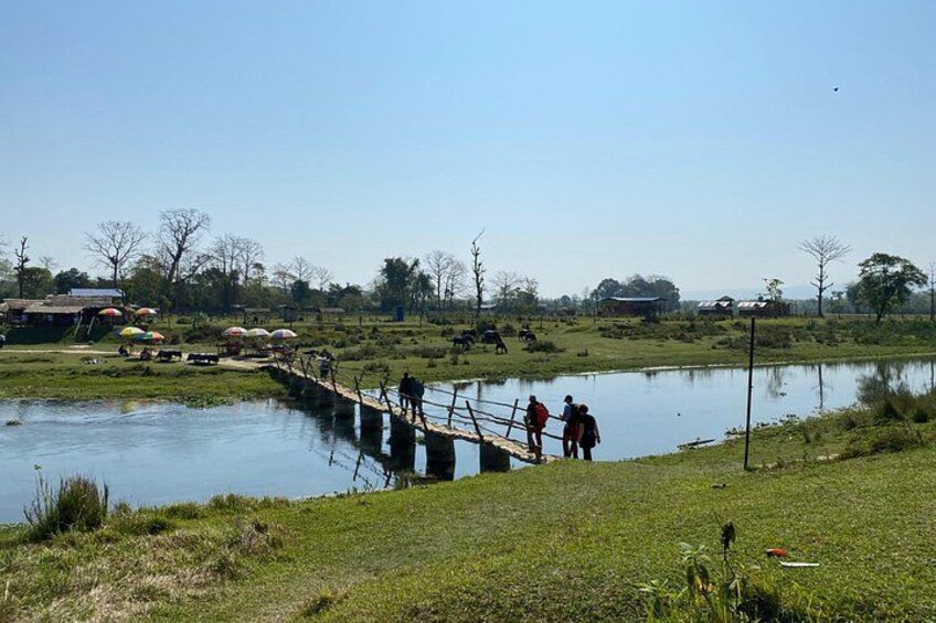 Eine Holzbrücke über einem ruhigen Fluss, Menschen gehen darauf, und im Hintergrund eine Wiese mit Bäumen und Hütten. 4