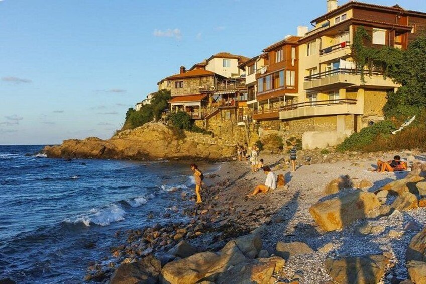 En klippefyldt strand med folk, der solbader og går, samt bygninger på en klippe med udsigt over havet. 5