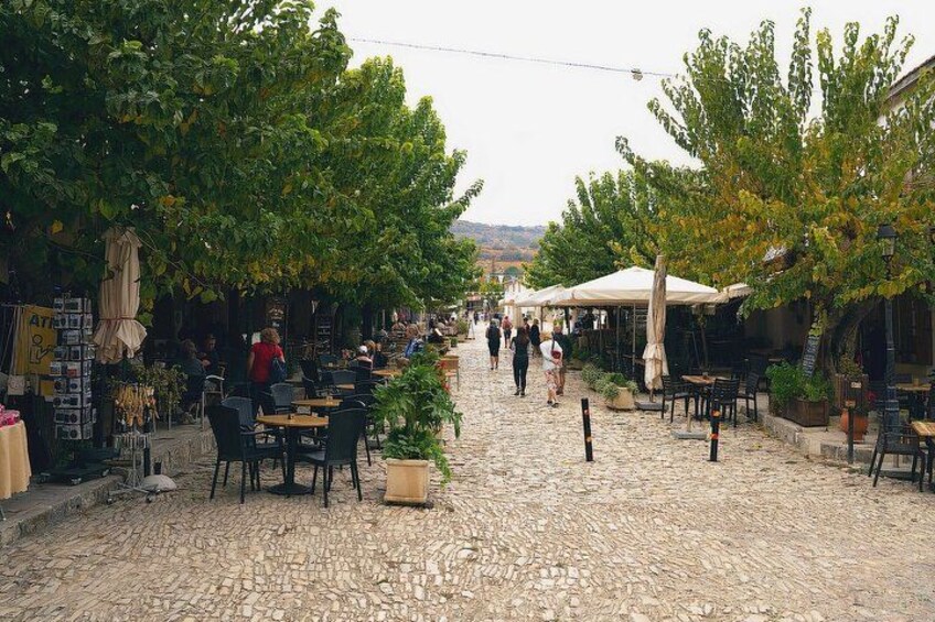 A cobblestone street lined with outdoor cafes and shops, trees, and a distant view of hills. 3