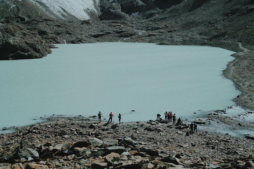 A group of people standing on a rocky shore by a calm lake surrounded by mountains. 1