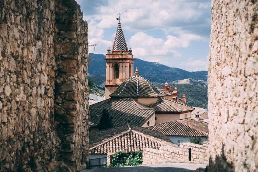 A historic town with a prominent church tower, terracotta-roofed buildings, and a mountainous backdrop. 2