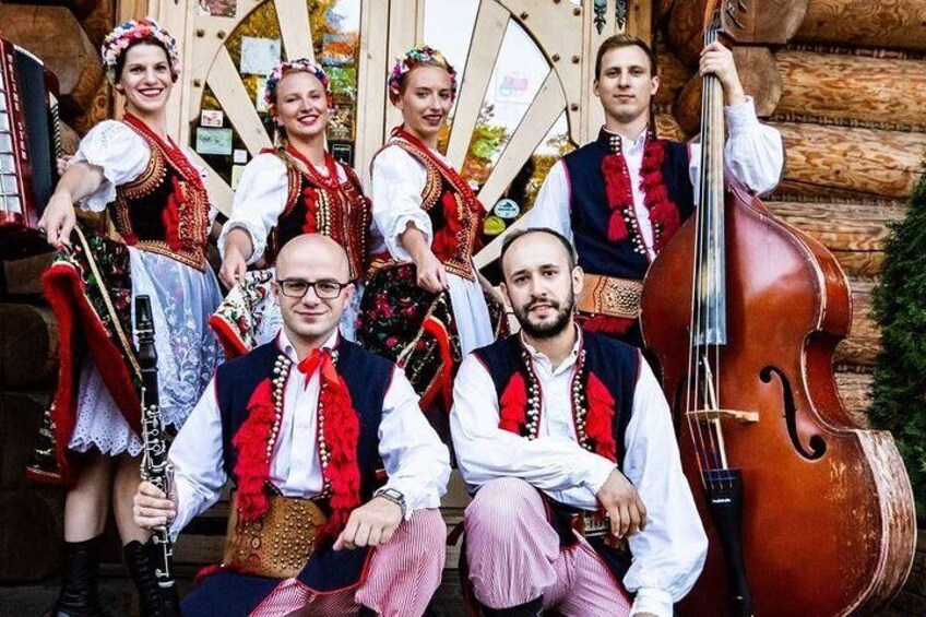 Un grupo de personas con vestimenta tradicional y instrumentos musicales, incluyendo un contrabajo, frente a una estructura de madera. 5