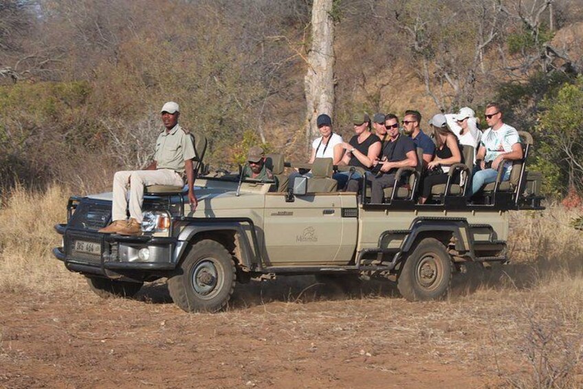 A safari vehicle with passengers, surrounded by dry grassland and sparse trees. 1