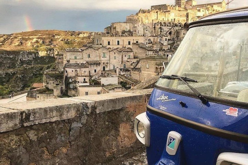 Un coche azul aparcado sobre un muro de piedra con vistas a un pueblo histórico, con edificios y un arcoíris en la distancia. 1