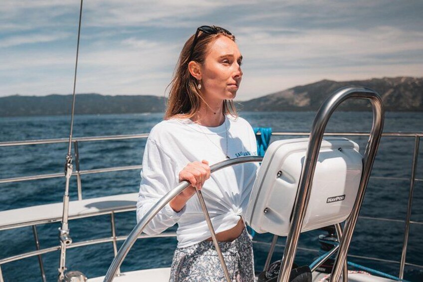 En person på en segelbåt, som håller i rodret, med en vattenyta och ett avlägset land i bakgrunden. 5