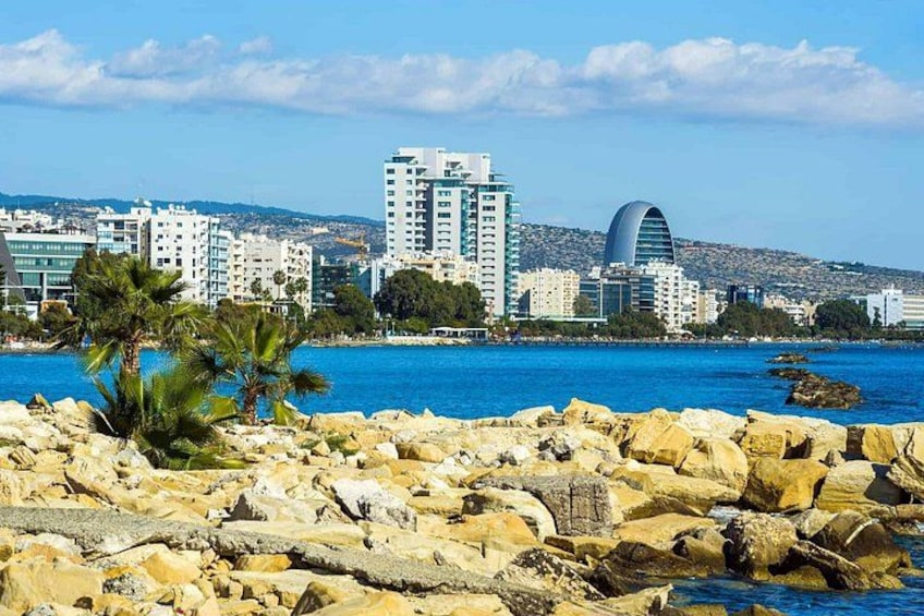 A coastal cityscape with rocky shoreline, palm trees, and modern buildings including a distinctive curved structure. 1