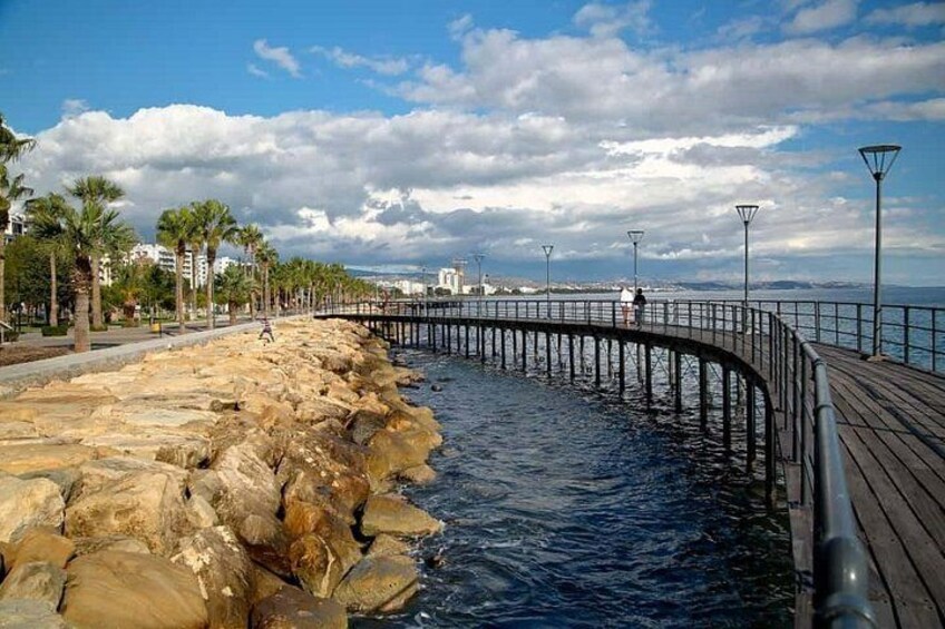 A coastal walkway with a wooden deck, rocky shoreline, palm trees, and a distant cityscape. 2