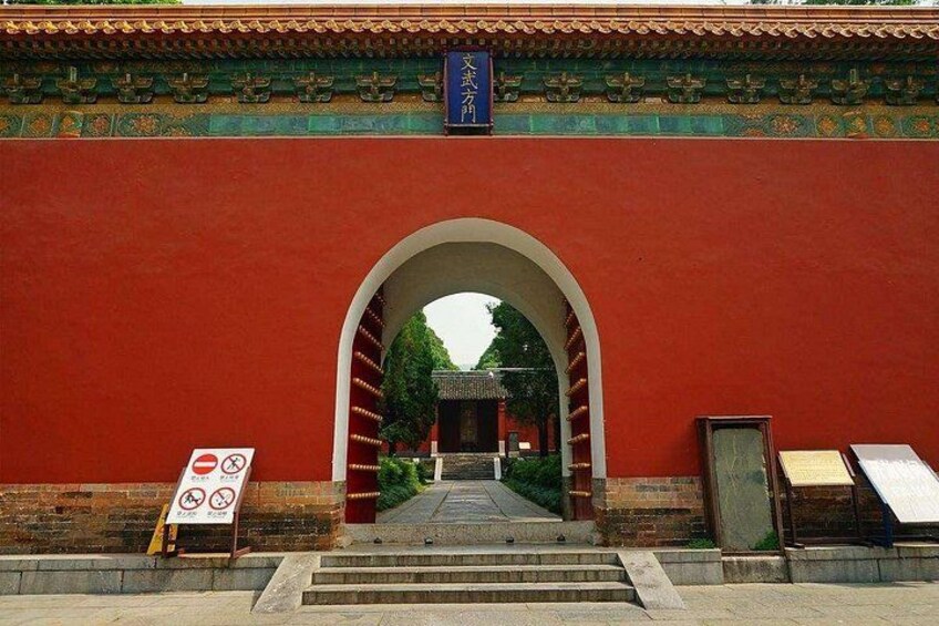 A red wall with an archway leading to a traditional building. There are signs with symbols and text on the wall. 4