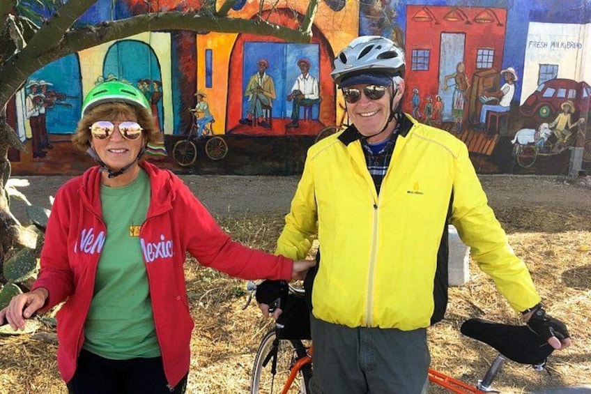 Two people standing in front of a colorful mural, one wearing a red jacket and the other in a yellow jacket. 1