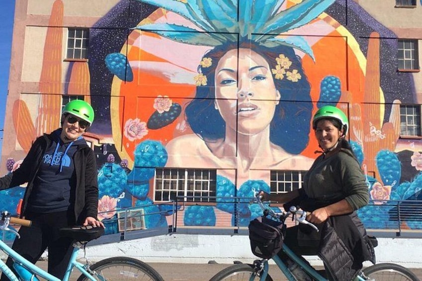 Two people on bicycles in front of a mural with a woman's face and cacti. 3
