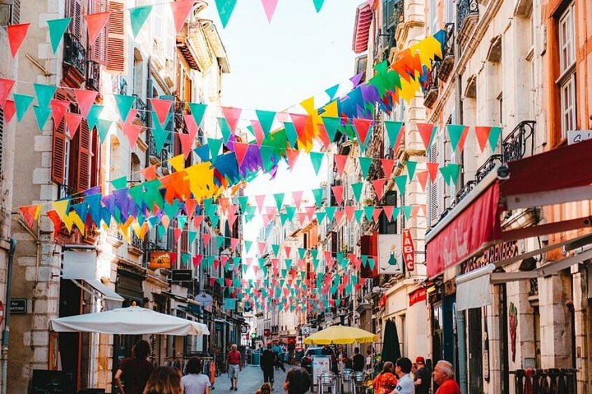A narrow street with colorful triangular flags strung overhead, buildings with balconies, and people walking. 1
