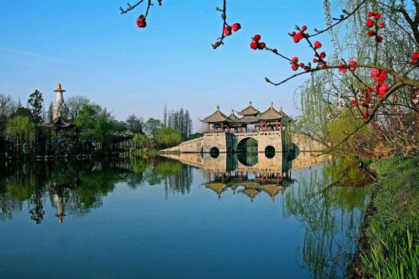 A traditional Chinese pavilion over a calm lake, surrounded by lush greenery and red flowering trees. 1