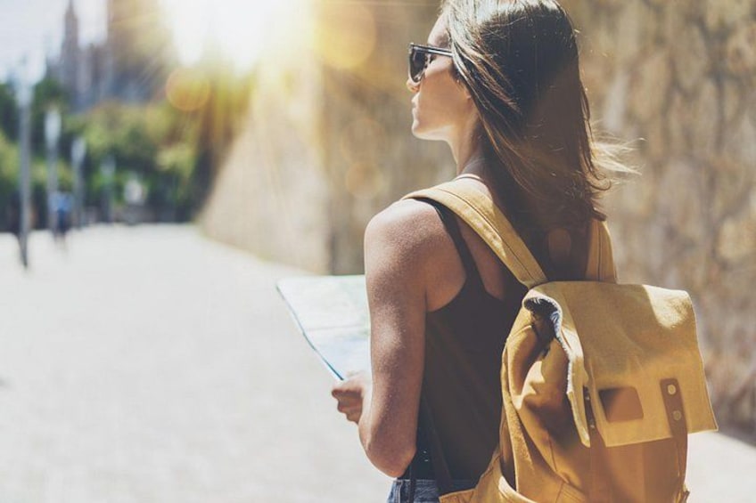 Eine Person hält eine Karte, trägt einen gelben Rucksack und geht auf einer sonnigen Straße. 5