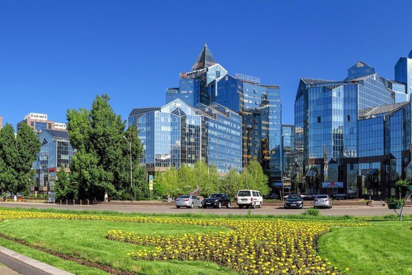 A cityscape with modern glass buildings, a green park with yellow flowers, and a clear blue sky. 1