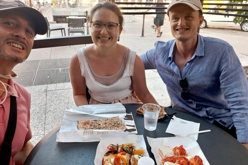 Drie personen zitten aan een buitentafel met eten en drinken. 3
