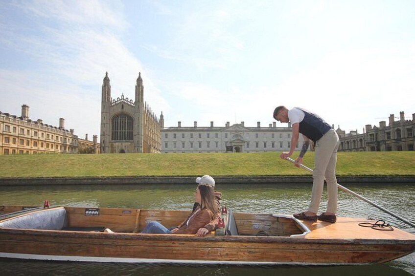 A wooden boat on a river with a person rowing, a woman sitting, and a historic building in the background. 4