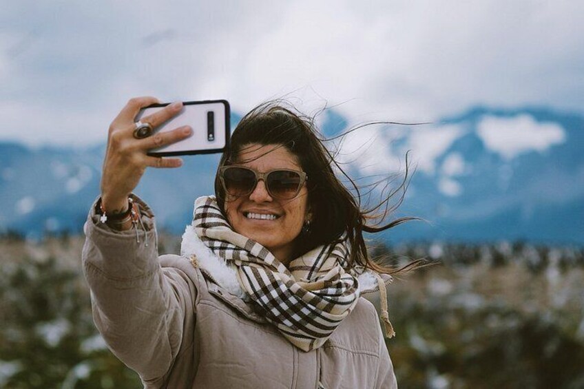 A person taking a selfie with a smartphone, wearing a scarf, and a jacket, with a mountainous landscape in the background. 4