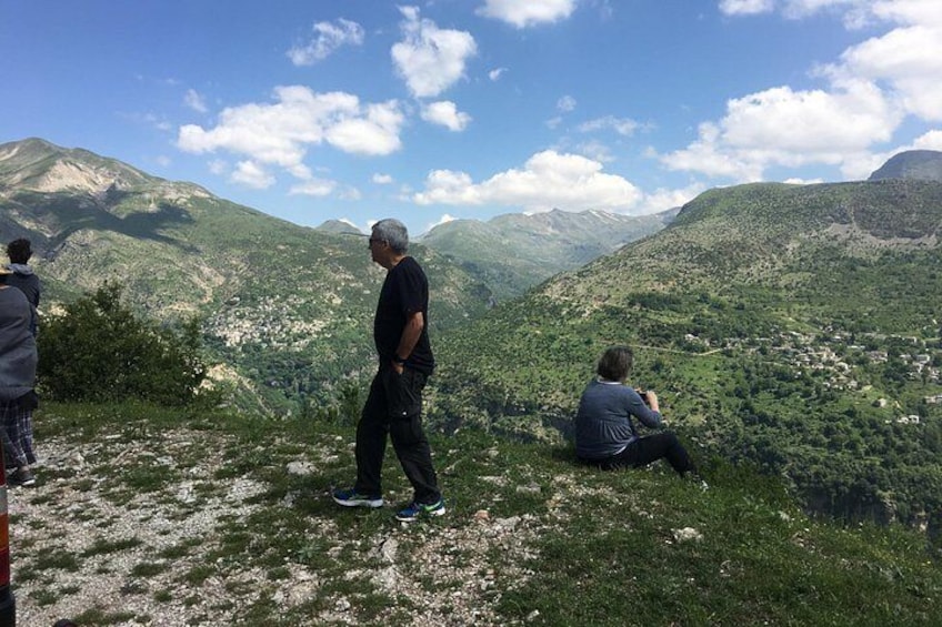 A mountainous landscape with a person standing and another sitting, overlooking a valley with scattered buildings. 3