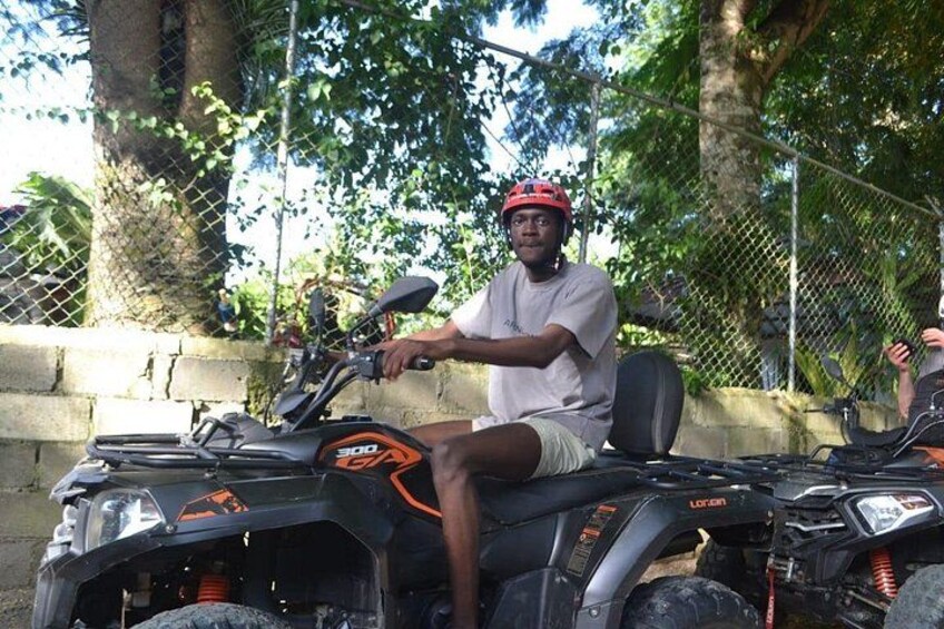A person sitting on a black ATV with orange accents, wearing a red helmet, and holding onto the handlebars. 3
